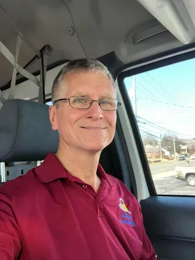 Man in maroon polo shirt wearing glasses, smiling inside a vehicle